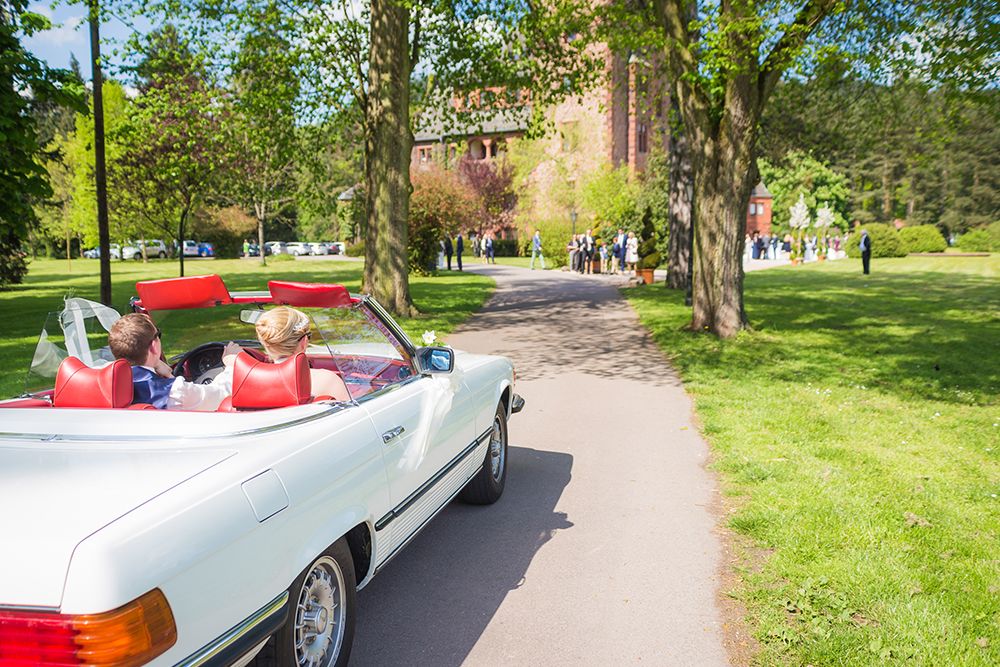 Ein Paar in einem weißen Cabrio mit roten Sitzen fährt einen von Bäumen gesäumten Weg entlang auf eine Gruppe von Menschen zu, die sich auf einer Wiese versammelt haben, und schafft so die perfekte Szene für eine märchenhafte Traumhochzeit.