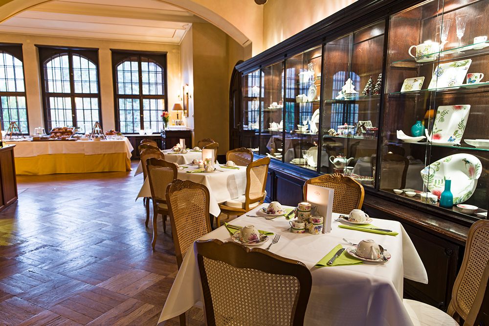 Ein gemütlicher Essbereich mit gedeckten Tischen für ein köstliches Frühstücksbuffet mit Teetassen und Tellern. Eine Glasvitrine stellt dekoratives Porzellan aus, während große Fenster im Hintergrund natürliches Licht hereinlassen und das Aroma von frisch gebrühtem Kaffee und verschiedenen Köstlichkeiten verstärken.