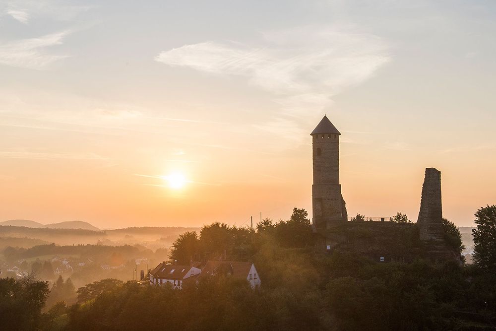Old stone tower and ruins on a hill with trees, houses and a glowing sunset in the background.