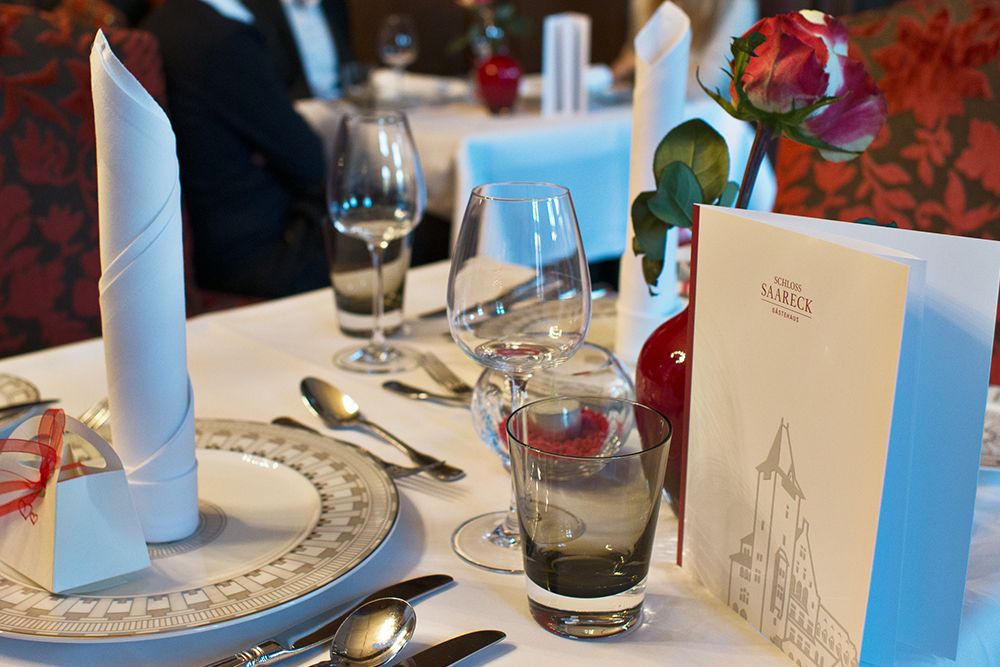 Elegant gedeckter Esstisch in einem großen Esszimmer mit gefalteten Servietten, Gläsern, einer Rose in einer Vase und einer Speisekarte mit einer Gebäudeillustration.