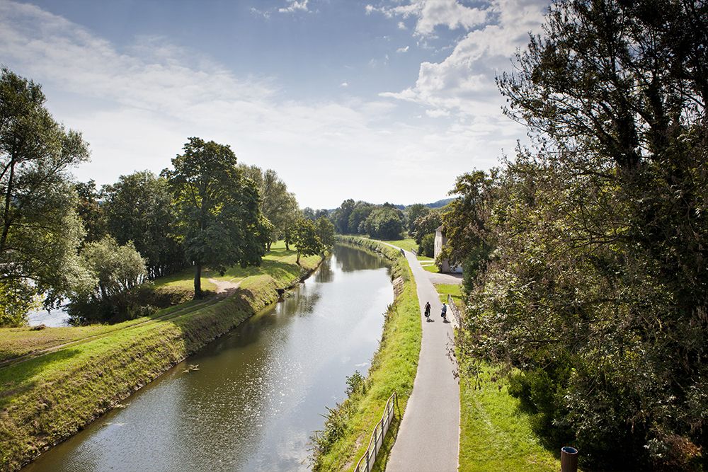 Ein Weg entlang eines Kanals mit zwei Radfahrern, umgeben von Bäumen unter einem teilweise bewölkten Himmel, erinnert an die ruhige Landschaft der saarländischen Traumschleifen, die für ihre Premium-Wanderwege bekannt sind.