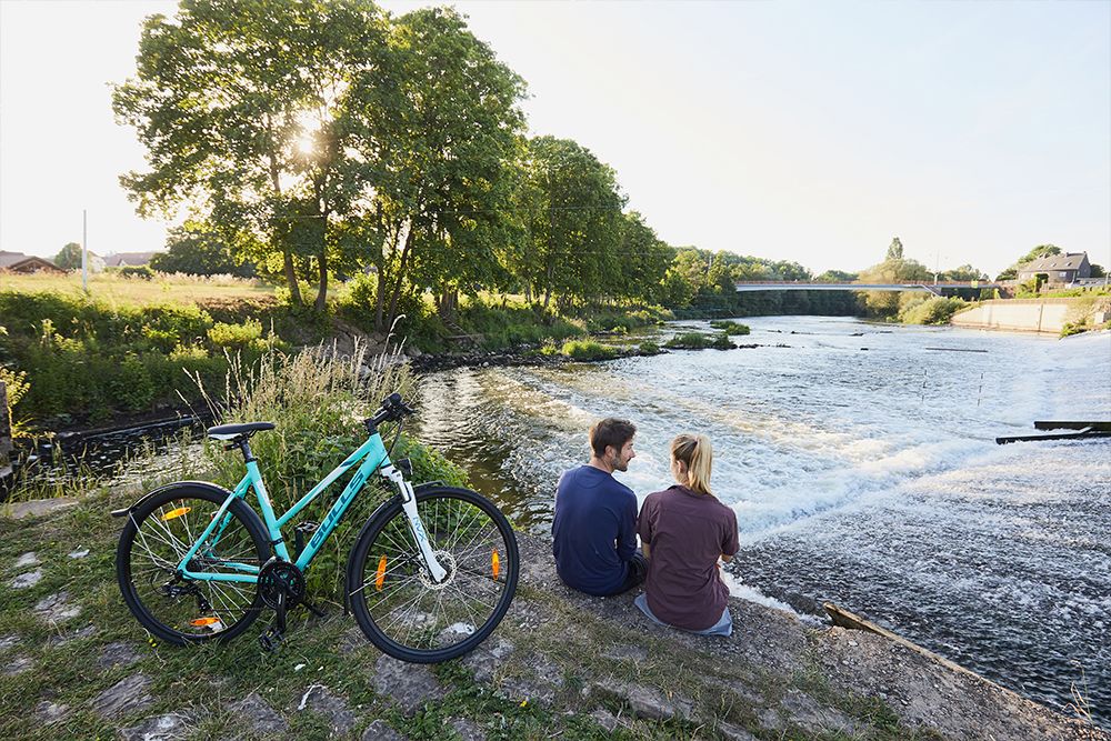 Ein Paar sitzt am Flussufer in der Nähe eines kleinen Wasserfalls, ihr blaues Fahrrad steht in der Nähe. Sie genießen die ruhige Schönheit der saarländischen Traumschleifen, mit Bäumen und einer Brücke, die die perfekte Kulisse für diesen idyllischen Ausflug auf diesen erstklassigen Wanderwegen bilden.