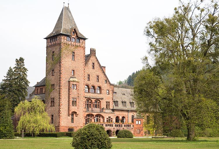 Schloss Saareck, ein großes, schlossähnliches Gebäude aus rotem Backstein mit einem hohen Turm und einem Satteldach, liegt elegant umgeben von üppigen grünen Bäumen und einem gepflegten Rasen und bietet ein zauberhaftes Hotelerlebnis nahe der Saar.
