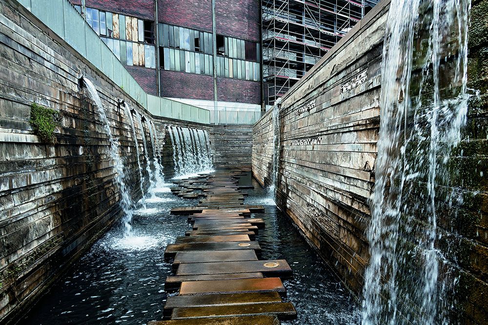 A narrow path with stepping stones crosses a water feature with cascading walls and industrial buildings in the background.