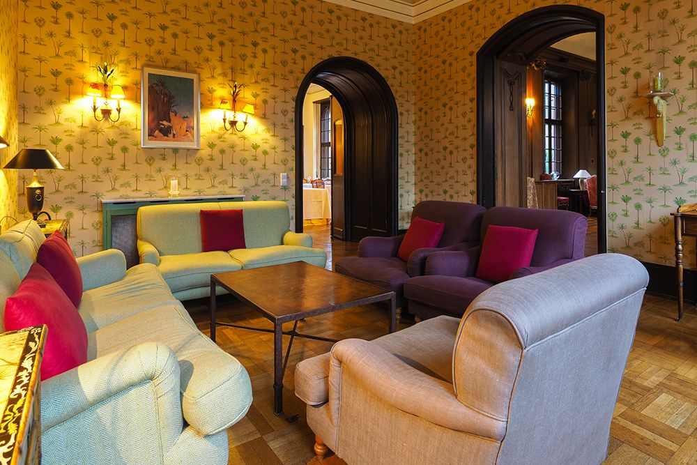 Cozy living room with patterned wallpaper, two sofas, armchairs and a coffee table. Wall lights brighten the room. Arched doors lead to a conservatory for relaxing and unwinding in nature.