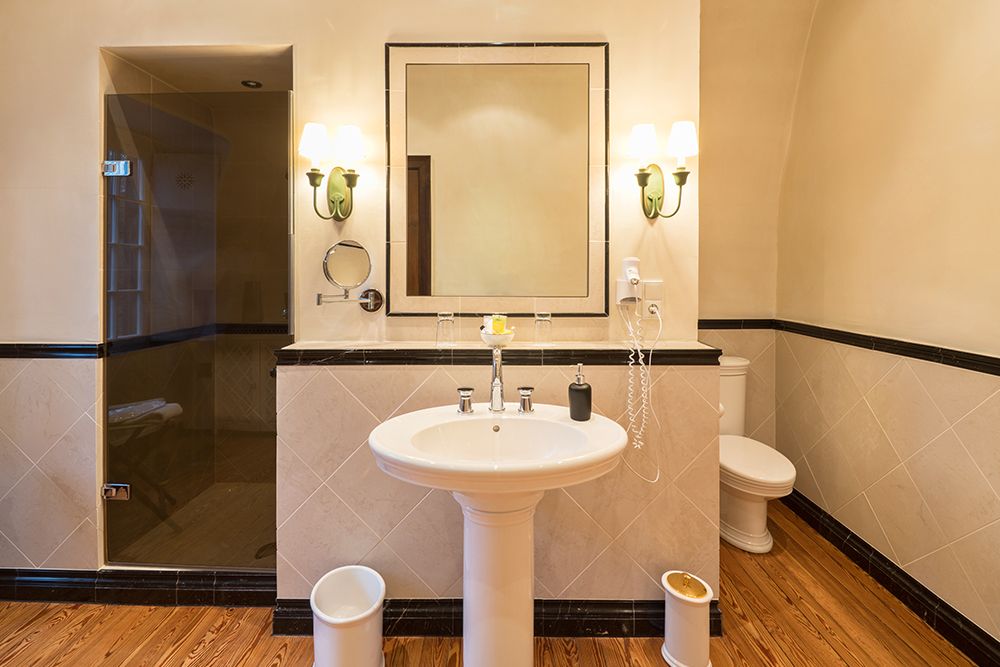 Badezimmer mit Standwaschbecken, quadratischem Spiegel, Wandleuchten, Dusche mit Glastür, Toilette und Holzboden.