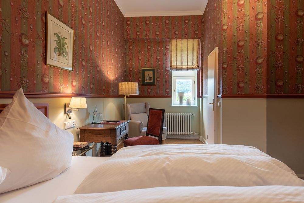Gemütliches Schlafzimmer mit Blumentapete, Doppelbett, Holzschreibtisch, Sessel, Fenster und gerahmter Wandkunst.