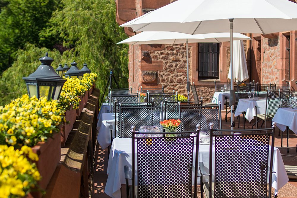 Außenterrasse mit leeren, mit weißen Tüchern bedeckten Tischen, umgeben von gelben Blumen und schwarzen Laternen. Weiße Sonnenschirme spenden Schatten auf der malerischen Schlossterrasse. Ein charmantes Backsteingebäude und üppiges Grün vervollständigen die ruhige Umgebung inmitten der Schönheit des Saarparks.