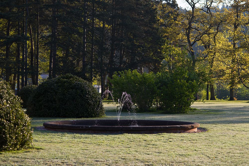 Ein kleiner Brunnen in der Mitte eines runden Beckens, umgeben von Gras und Bäumen in einer Parkanlage.