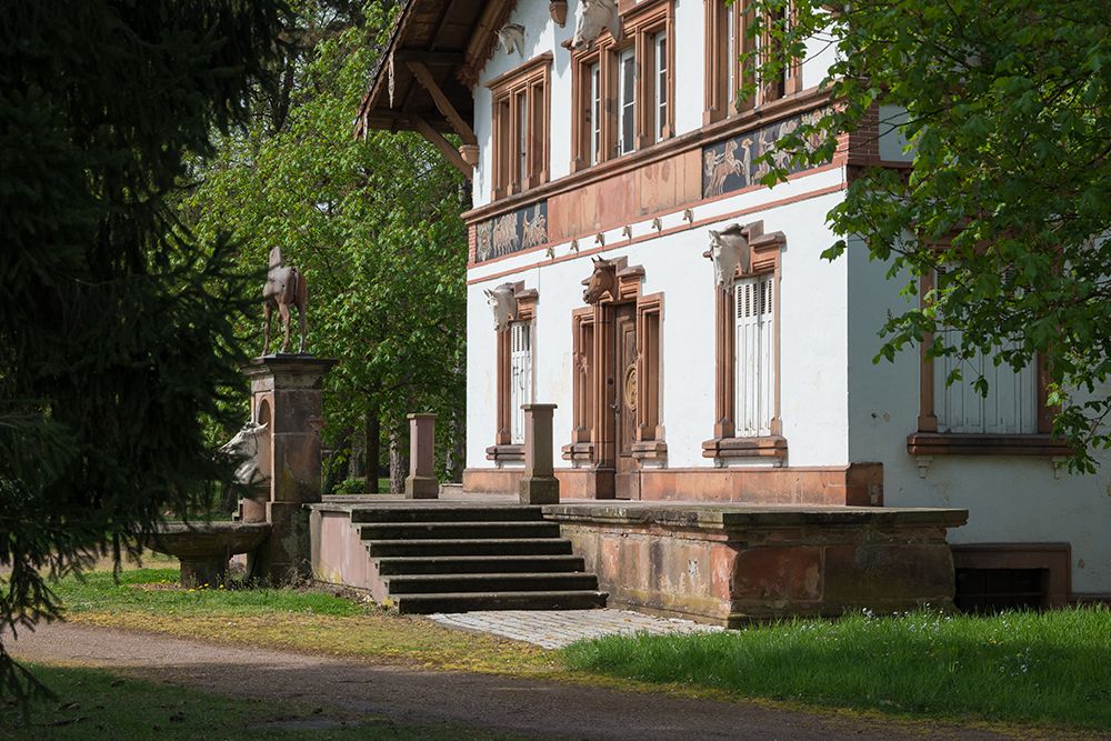 Ein historisches Gebäude mit Steintreppen, die zu einer Holztür führen, umgeben von grünen Bäumen. Neben dem Eingang ist eine Skulptur zu sehen.