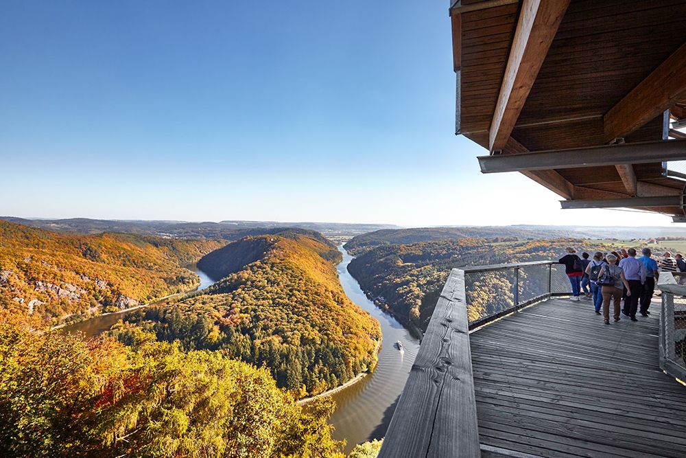 The observation deck, ideal for day trips, offers stunning views of a curving river surrounded by forested hills on a clear day. A group of people enjoy the scenery just a short distance from the hotel.