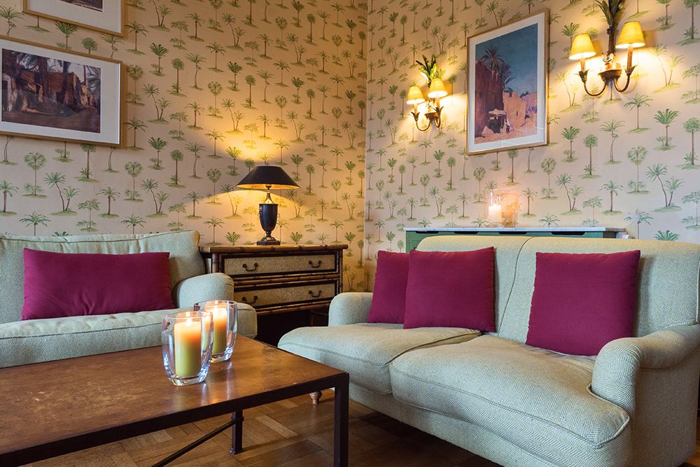 A living room with green sofas, pink cushions and a wooden table with candles exudes relaxation. The walls are decorated with palm wallpaper and framed artwork, while two wall lamps spread a warm glow.