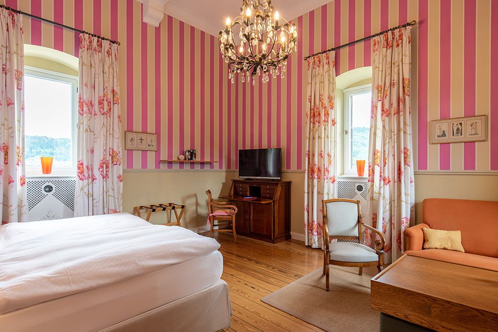 Ein Schlafzimmer mit rosa gestreiften Wänden, Kronleuchter, Doppelbett, Fernseher, orangefarbenem Sofa, zwei Fenstern mit geblümten Vorhängen und Holzboden.