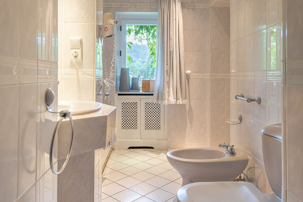 Gefliestes Badezimmer mit Waschbecken, Bidet, Toilette und Fenster mit Vorhängen. Durch das Fenster fällt natürliches Licht ein und erhellt den Raum.