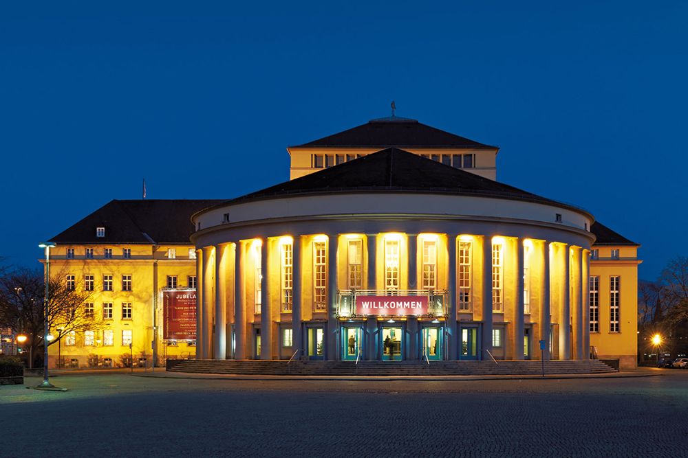 Ein Theatergebäude bei Nacht, dessen Säulen von hellen Lichtern erhellt werden und über dem Eingang ein Schild mit der Aufschrift „Willkommen“ steht, lädt die Gäste ein, in einen kulturellen Abend einzutauchen, in dessen historischen Sälen die Harmonie der Kammermusiktage erklingt.