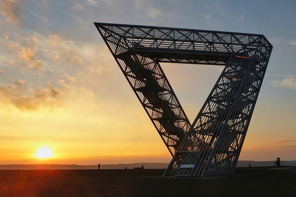 Triangular metal sculpture set against the backdrop of a sunset with clear skies and a distant horizon - a breathtaking sight for anyone exploring the famous sights of Saarland.