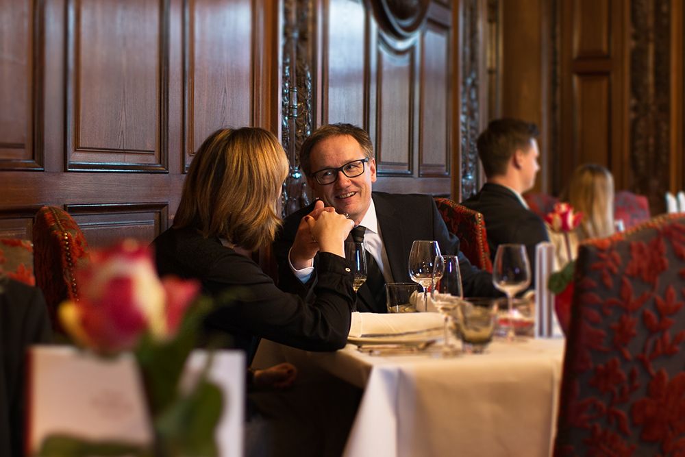 Im Schloss Saareck sitzen zwei Personen in formeller Kleidung an einem Restauranttisch, halten sich an den Händen und lächeln einander an. Die Kulisse ist mit kunstvollen Holztäfelungen und elegantem Dekor ausgestattet und verkörpert eine einmalig schöne Weise, die diesem Moment das Gefühl eines Festes verleiht.