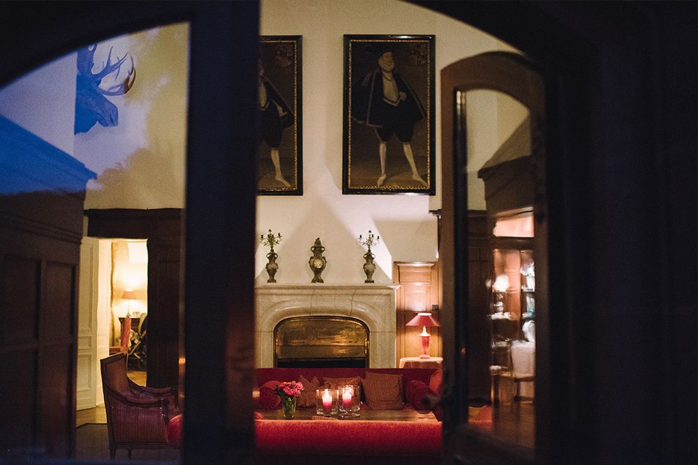 Gemütliche Lobby mit Kamin, roten Sofas und Kerzen, die ein warmes Licht verbreiten. Gerahmte Porträts hängen darüber und verleihen Charme. Holztüren rahmen die Aussicht ein, perfekt für einen Aperitif in der schwach beleuchteten Atmosphäre.