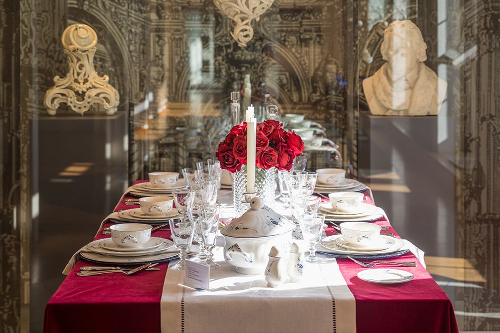 Elegant dining table set with porcelain dishes, crystal glasses, red tablecloth and a centerpiece made of red roses, reminiscent of the artistic decoration of the great castles of Saarland.