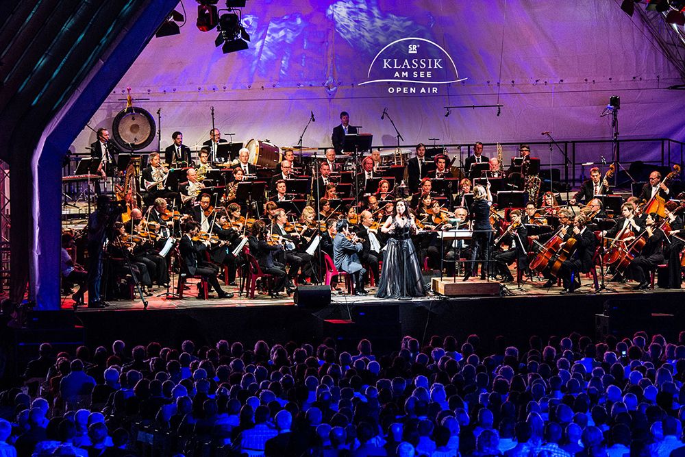 Beim zauberhaften Open-Air-Konzert „Klassik am See“ spielen Orchester und Dirigent unter dem Nachthimmel und verzaubern ein großes sitzendes Publikum mit wunderschöner Musik.