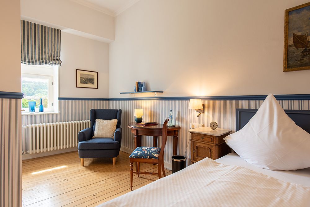 Gemütliches Schlafzimmer mit Einzelbett, Sessel am Fenster, kleinem Schreibtisch und dekorativer Lampe. Gestreifte Wände und Holzboden.