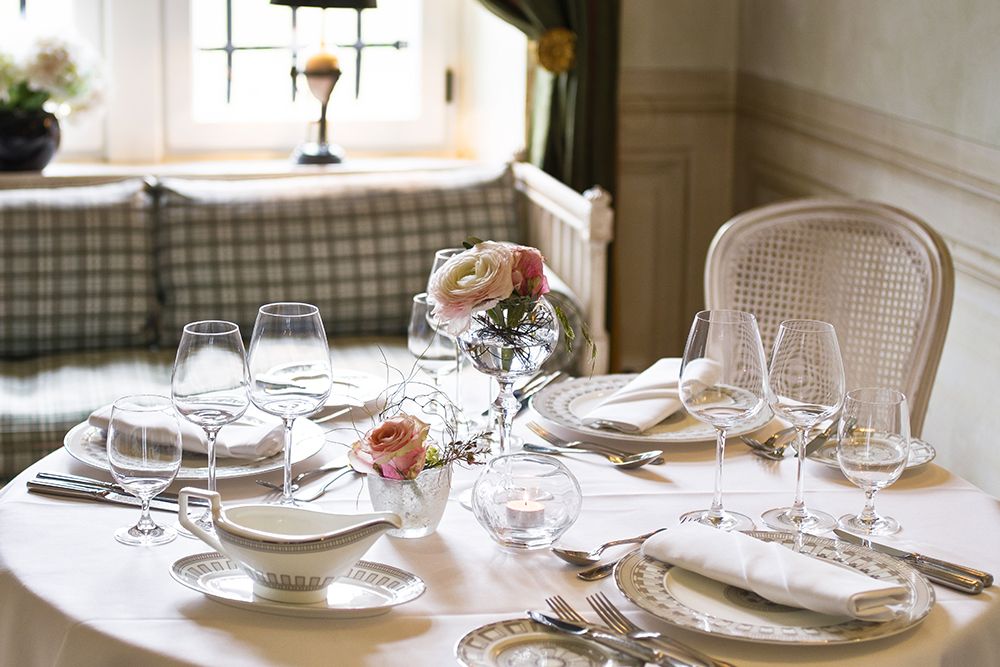 Elegant gedeckter Esstisch für ein Abendessen zu zweit, mit weißem Geschirr, Weingläsern, Stoffservietten und einem Blumengesteck in einem Raum mit einem karierten Fenstersitz, der eine intime Atmosphäre und perfekte Privatsphäre bietet.