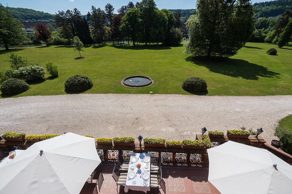Erhöhte Ansicht eines Gartens mit einem kleinen runden Brunnen, umgeben von Bäumen und Rasen. Im Vordergrund ist eine Terrasse mit einem gedeckten Tisch und zwei großen weißen Sonnenschirmen zu sehen.