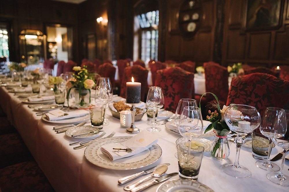 Elegant dining table with white tablecloth, glasses, cutlery, plates, flower arrangements and a dark candle in a wood paneled room with red upholstered chairs.