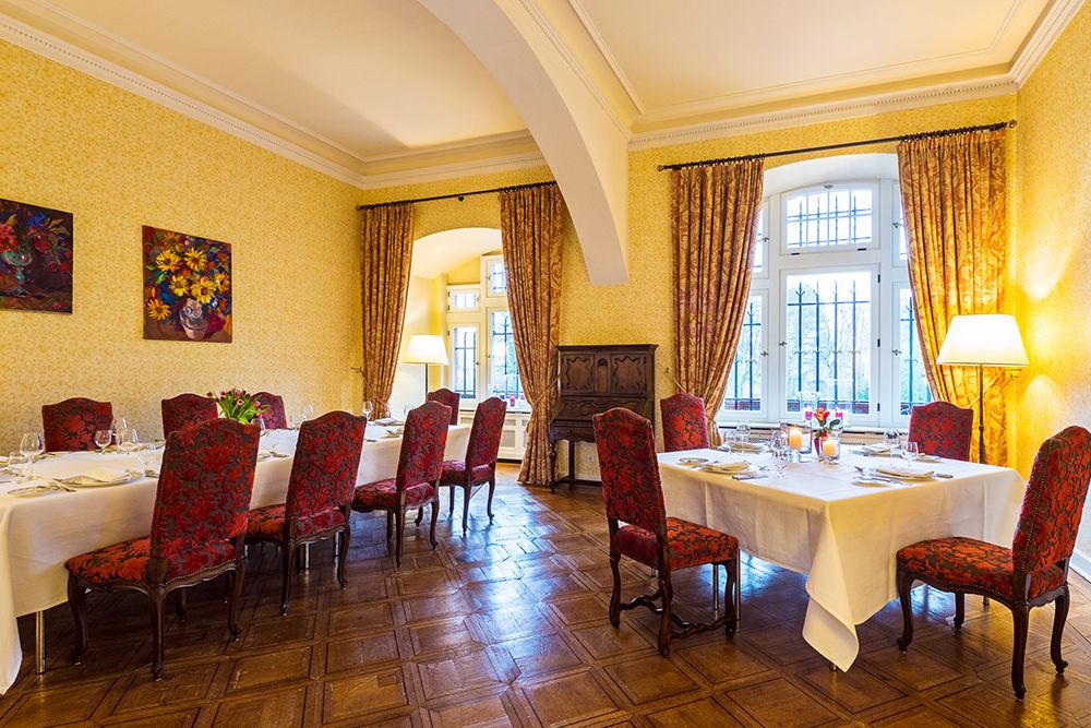 Elegant dining room with two set tables, red upholstered chairs and wooden floor. Perfect for meetings or celebrations, it features high windows with curtains, a sideboard decorated with lamps and floral paintings reminiscent of Anna Boch's delicate artistry.