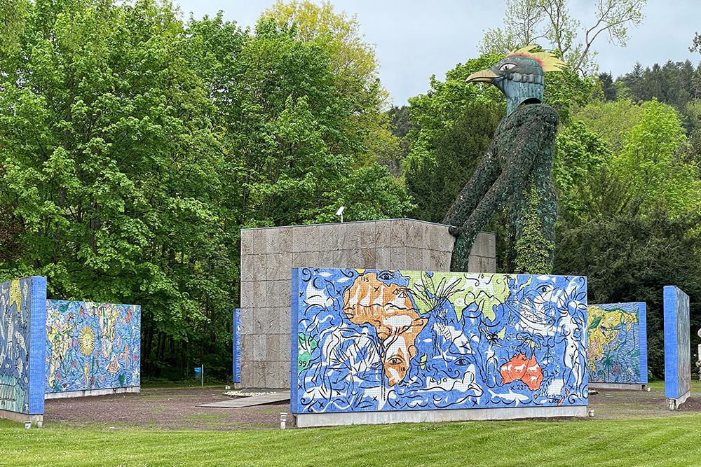 Large green bird-shaped sculpture next to a stone structure, surrounded by colourful mosaic tiles, in a lawn with trees.
