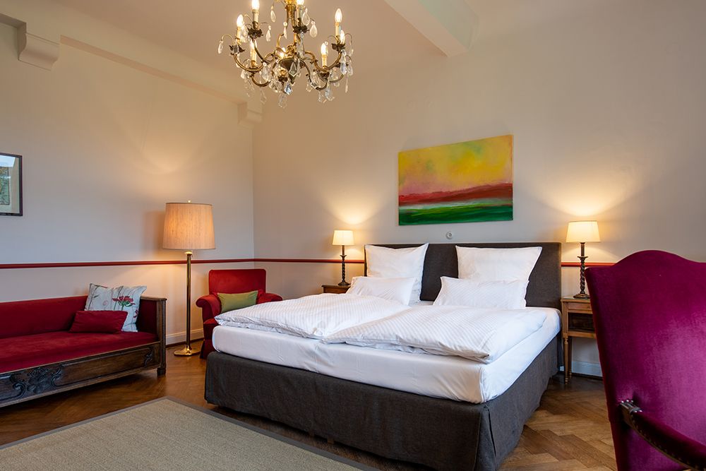 Ein ordentliches Schlafzimmer mit einem Doppelbett, zwei Nachttischlampen, einem Kronleuchter, Kunstwerken an der Wand und einem roten Loungesessel.