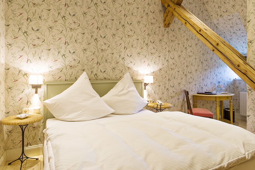 Ein gemütliches Schlafzimmer mit einem Doppelbett, geblümter Tapete, Holzbalken, zwei Nachttischen mit Lampen und einem kleinen Schreibtisch mit Stuhl.