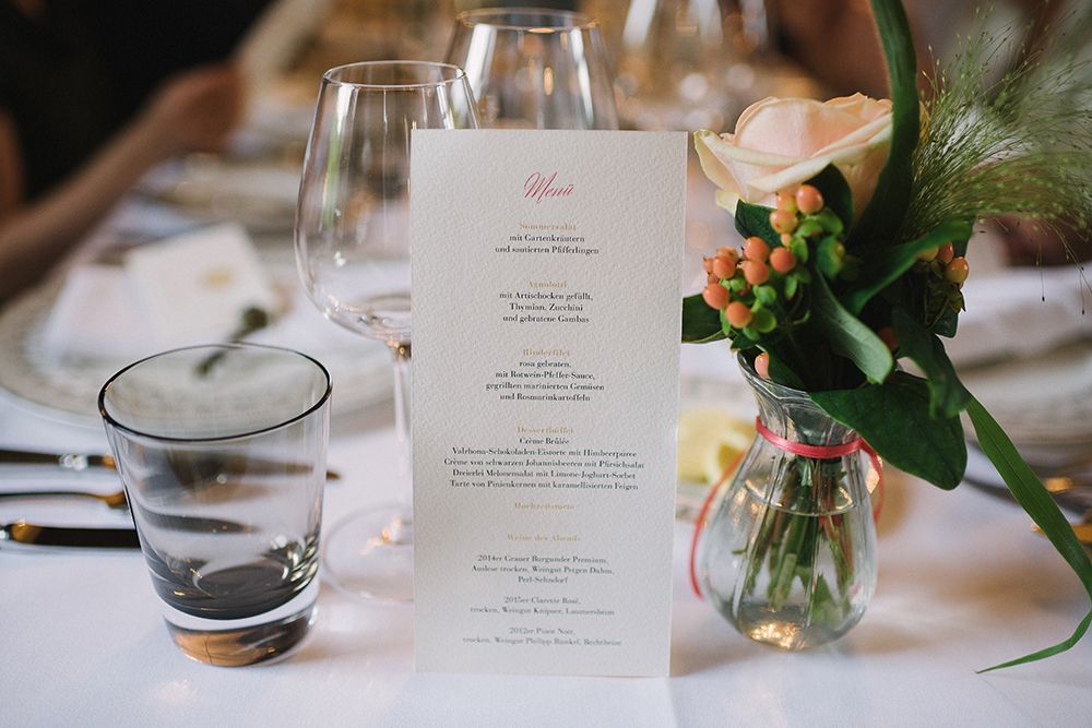 Ein Esstisch im Schloss Saareck ist elegant gedeckt mit einem Menü, Weingläsern, einem Glas Wasser und einer Vase mit einer rosa Rose und Beeren. Diese festliche Szene lädt die Gäste zum Feiern und Genießen ein.