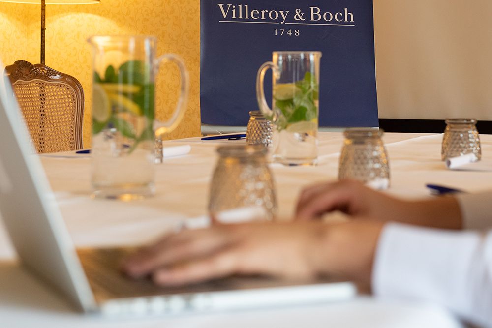 Hände tippen auf einem Laptop in einer Konferenzumgebung mit Krügen mit aromatisiertem Wasser auf einem Tisch und einem Banner von Villeroy & Boch im Hintergrund, das erstklassige Tagungsort-Räumlichkeiten präsentiert, die mit hochmoderner Tagungstechnik ausgestattet sind.