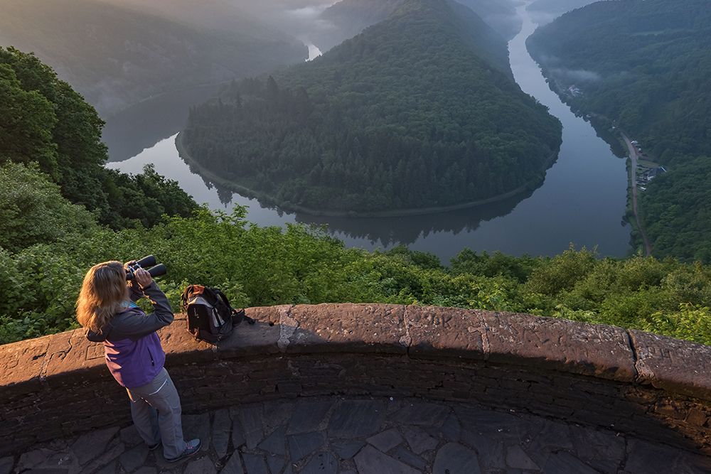 At sunrise, a person with a camera and a backpack enjoys the view from a stone lookout point, overlooking a bend in the river surrounded by lush greenery, reminiscent of the scenic beauty of Saarland's castle landmarks.
