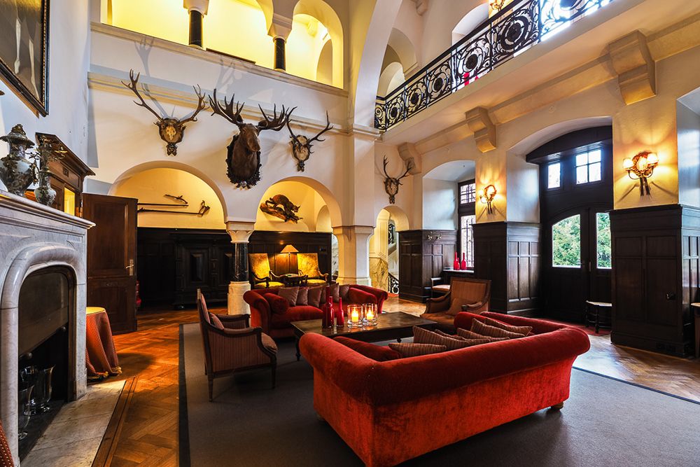 Das geräumige Wohnzimmer mit seinen hohen Decken erinnert mit seinen roten Sofas, dem Steinkamin und den aufgespießten Tierköpfen an die Lobby eines großen Hotels. Bei der dekorativen Beleuchtung können Sie einen Aperitif genießen und dabei die Treppe mit ihrem verzierten Geländer im oberen Stockwerk bewundern.