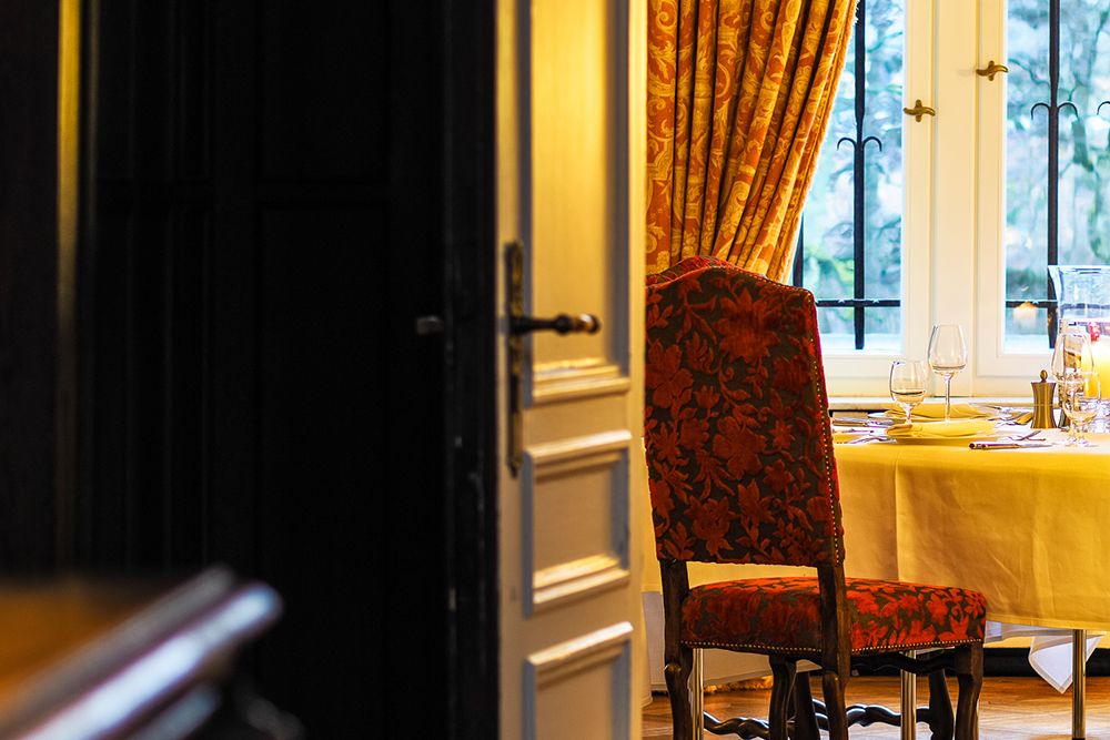 Reminiscent of the style of Anna Boch, the dining room is set for a meal with red patterned chairs, a yellow tablecloth and glassware. A partially open door frames the scene while curtains hang over the windows.