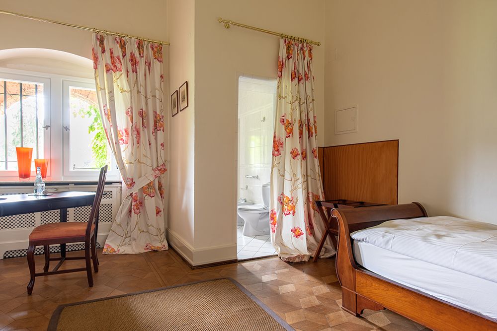 Einfaches Schlafzimmer mit Einzelbett, Schreibtisch und hellen Blumenvorhängen. Angrenzend führt eine offene Tür zum Badezimmer.