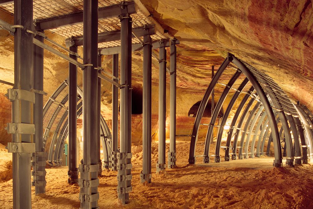 Underground mining tunnel with metal support arches and pillars, illuminated by warm light, revealing rock walls and sandy floor.