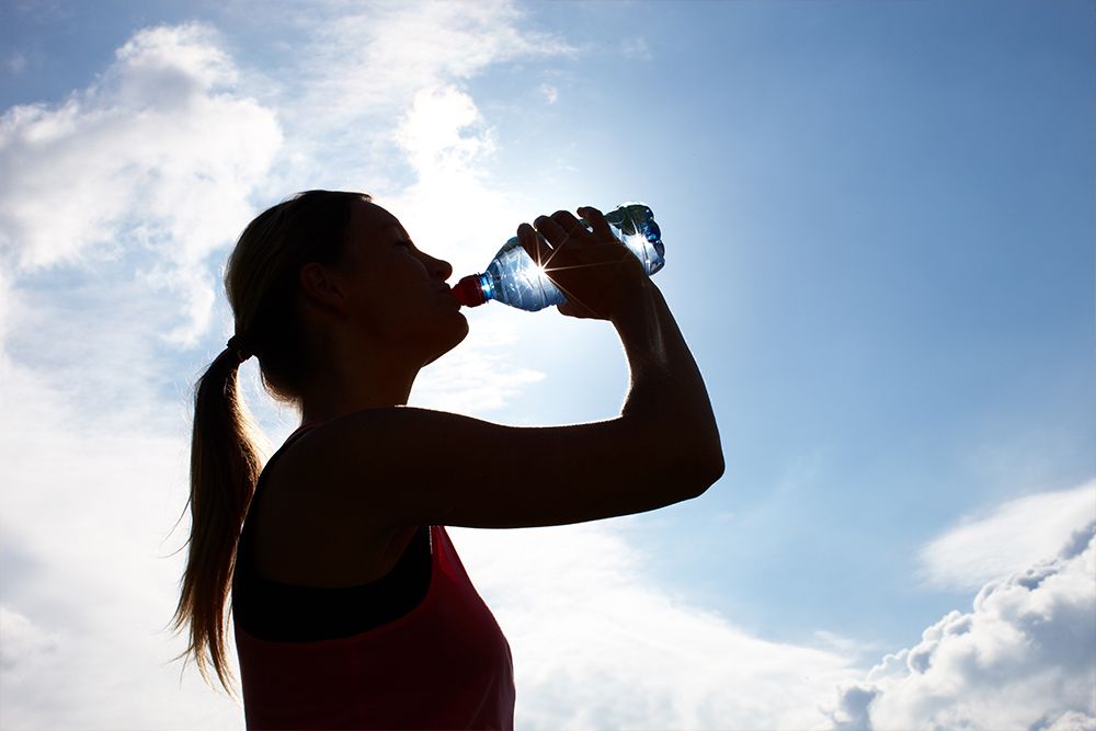 Silhouette of a person drinking a bottle of water after an invigorating jogging session against a bright sky with some clouds.