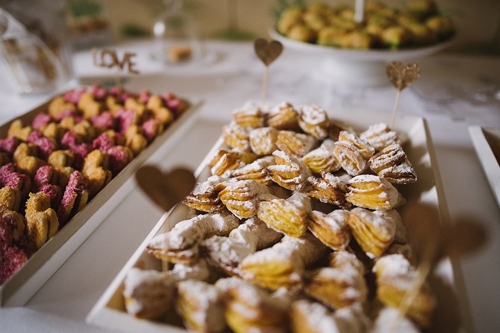 A tray of puff pastries dusted with powdered sugar, with a blurry tray of pink treats and a “LOVE” sign in the background.